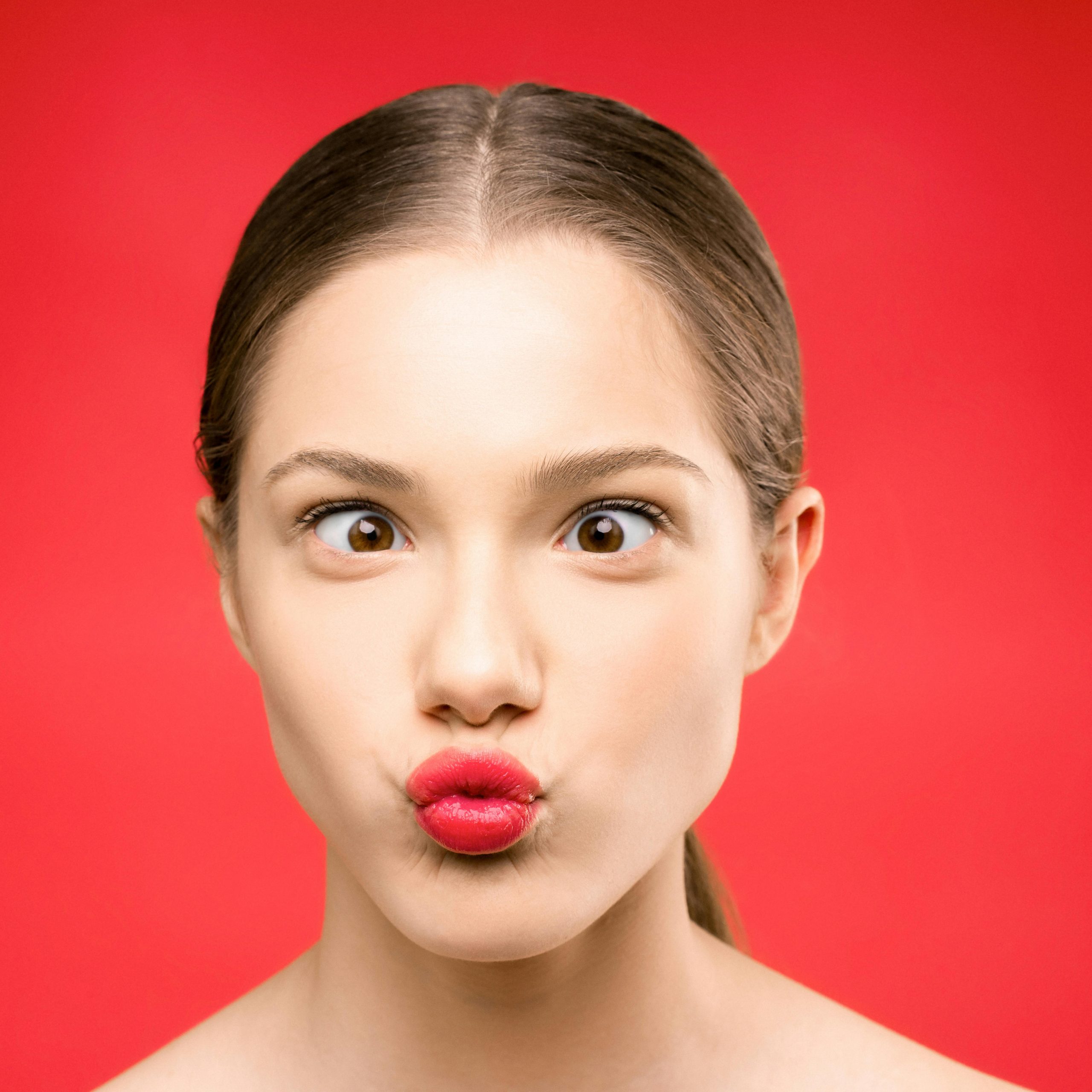 Young woman making a playful pout with bright red lipstick against a vibrant red background.