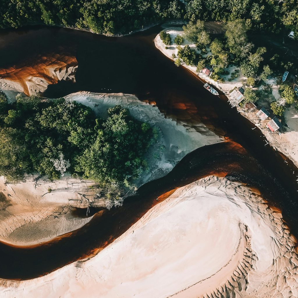 Breathtaking aerial view of boat moored on brown river flowing among sandy terrain near lush green forest in tropical countryside